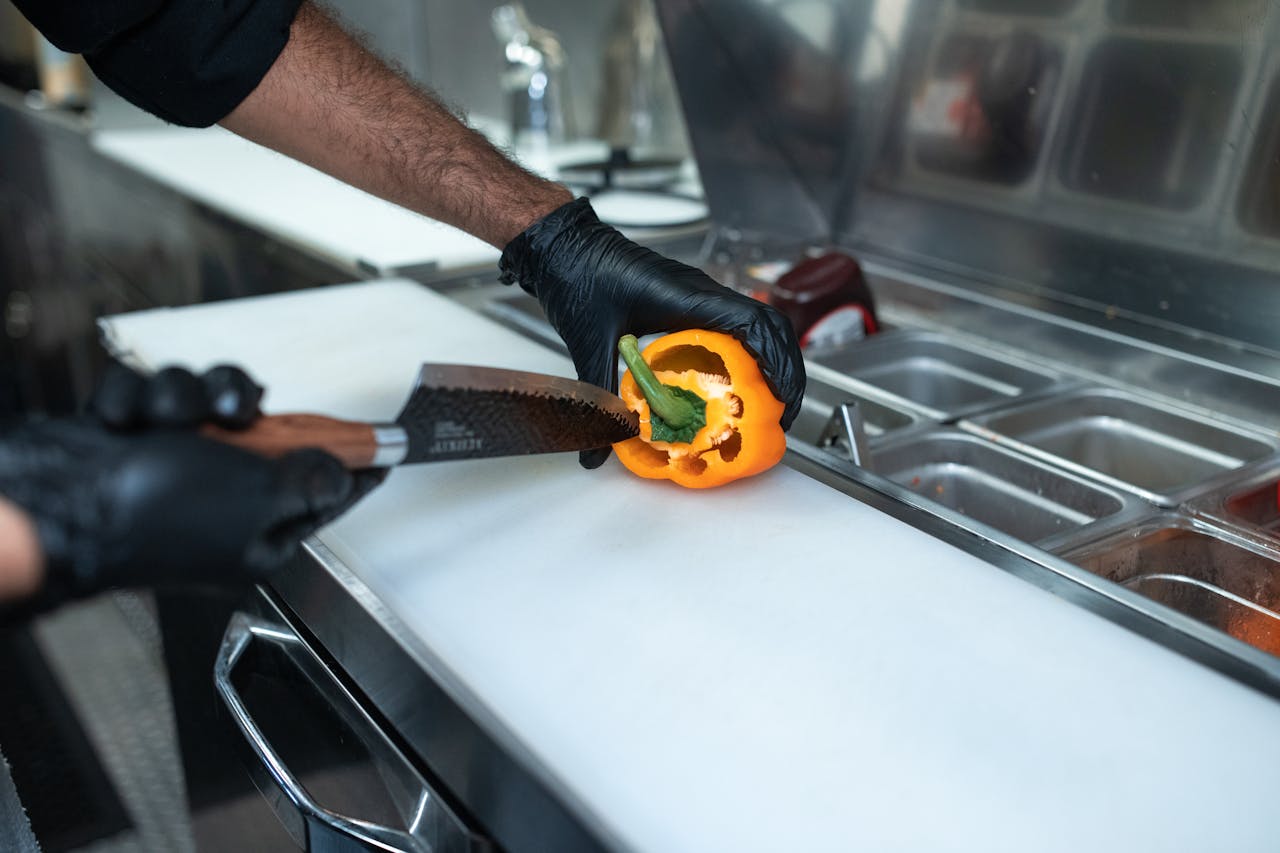 A chef in black gloves slices a yellow bell pepper on a cutting board in a commercial kitchen setting.