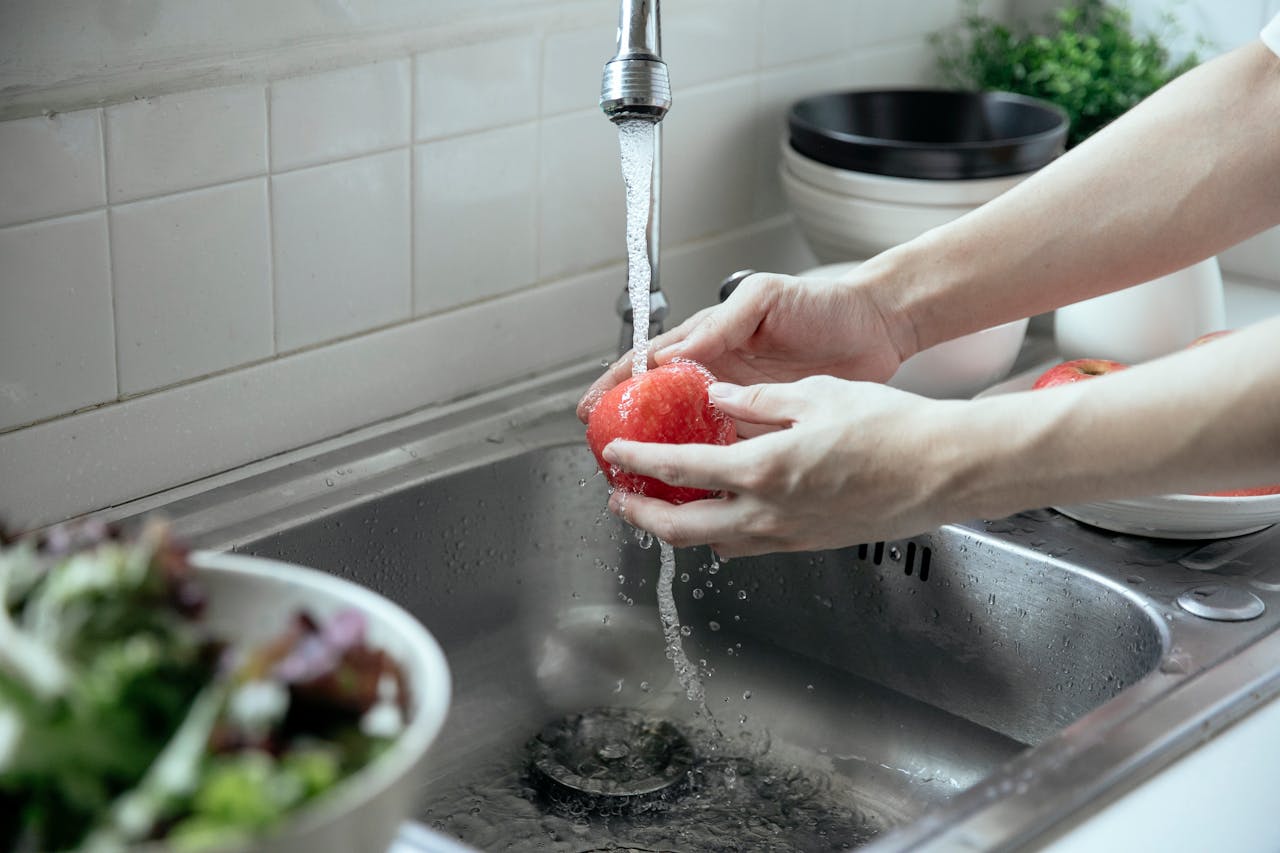 Close-up of hands washing a red apple under running tap water in a kitchen sink.
